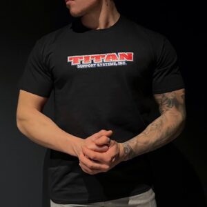 Lihaksikas mies mustassa Titan Support Systems -paidassa tummalla taustalla.